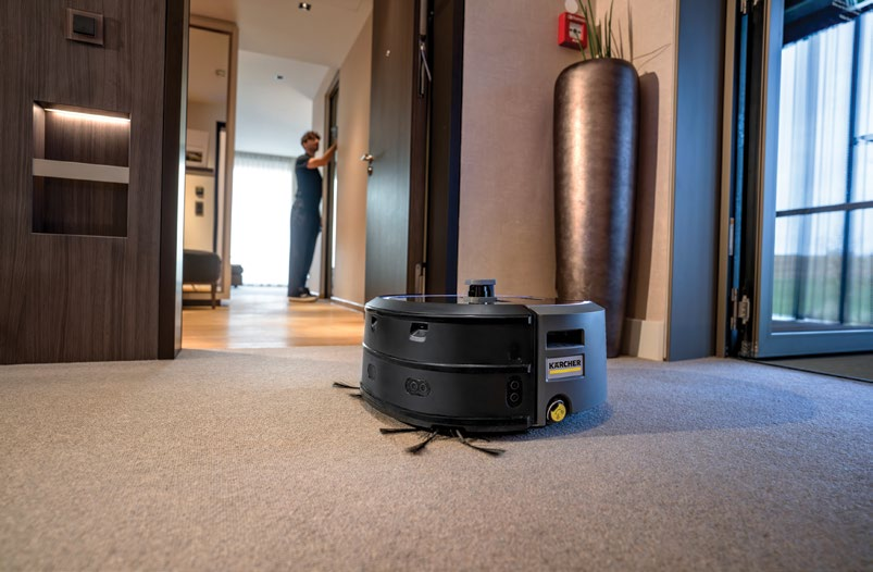 Kärcher KIRA serisi otonom temizlik robotu bir otel koridorunda halı zemini temizlerken, arka planda bir personel görevini sürdürüyor